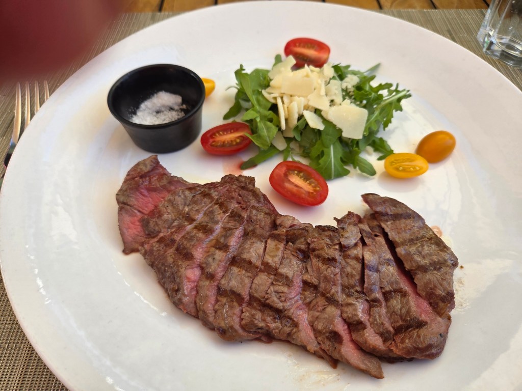 A plate featuring sliced grilled steak alongside a fresh salad with arugula, cherry tomatoes, and shaved cheese, served with a small bowl of salt.