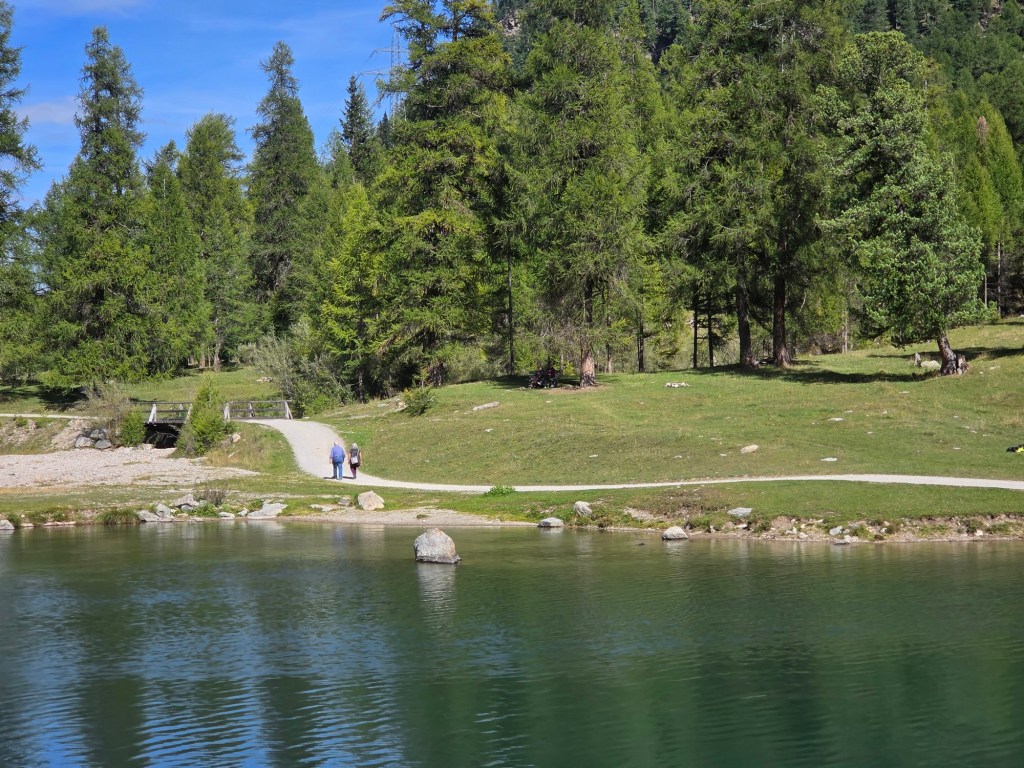 A serene landscape with a calm lake surrounded by lush green trees, featuring two people walking along a path by the water.